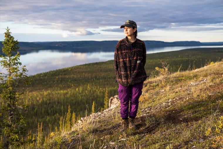 Kelly in ball cap and hiking dear looks from hilltop with large river and meadow behind her