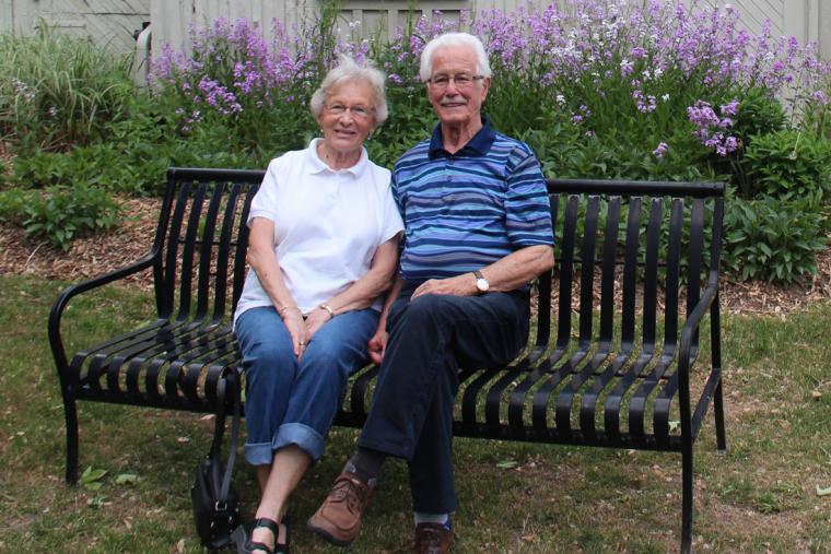 Barb and Joe Maxwell sit on black iron bench in front of garden of purple wild flowers