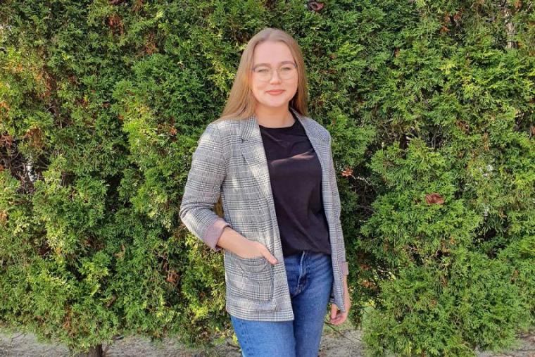 Priscilla in suit jacket smiles standing in front of cedar trees