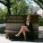 A woman with long hair dressed in heels and graduation gown sitting on a bench in front of a landscape architecture sign.