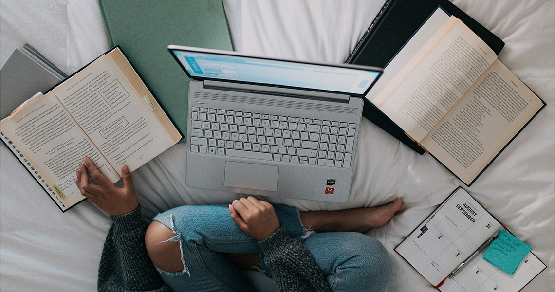 A person with crossed legs sitting in a bed with laptop and multiple books spread out in front of them.