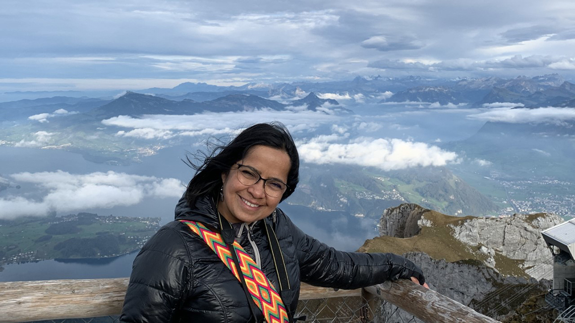 A woman in a black coat and glasses smiling, with large beautiful mountains in the background.