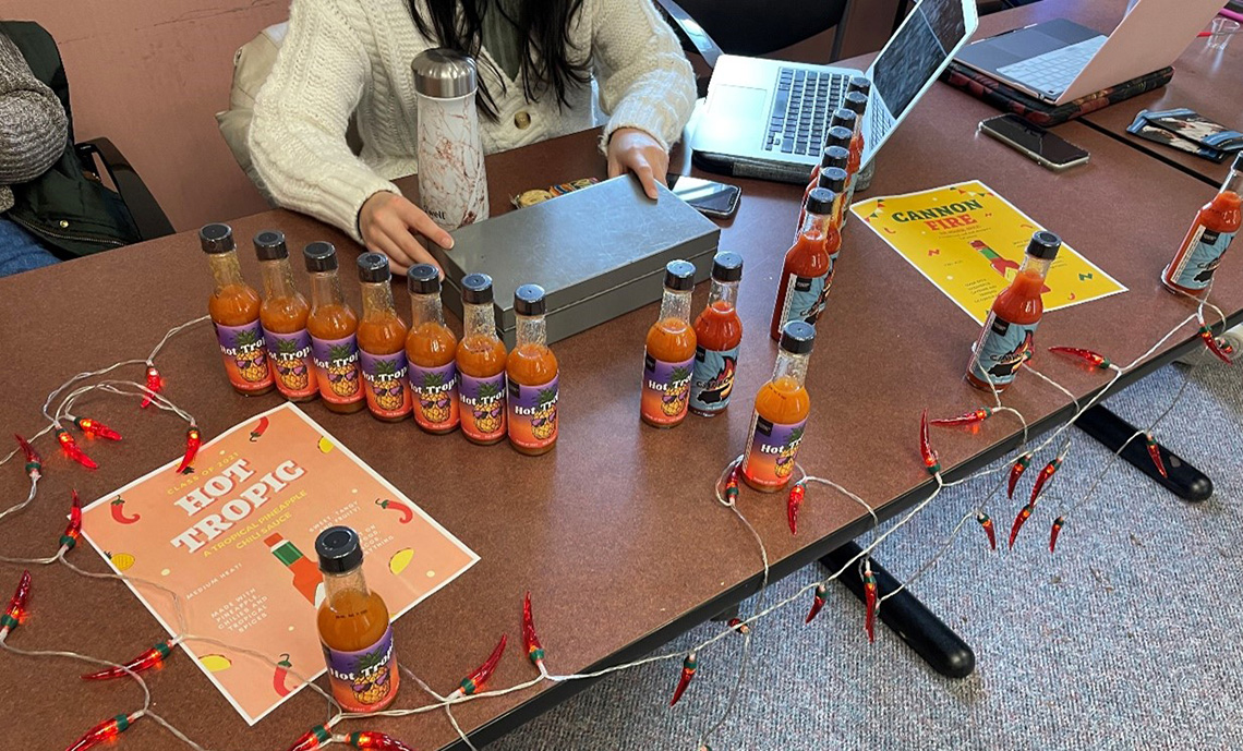 A student sitting at a table selling bottles of hot sauce.