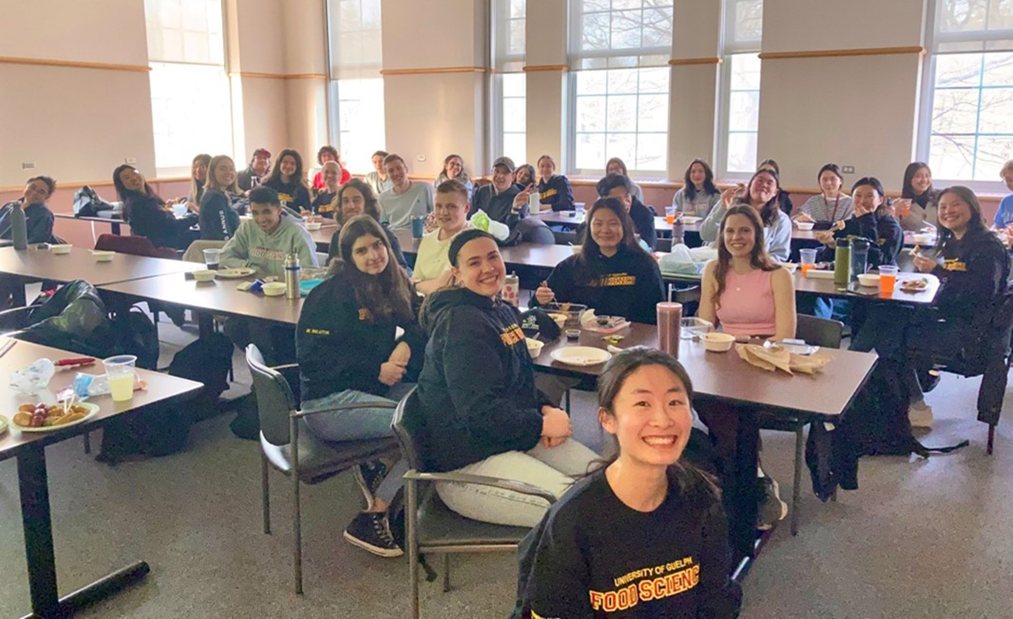 A large group of student eating lunch and smiling in a classroom.