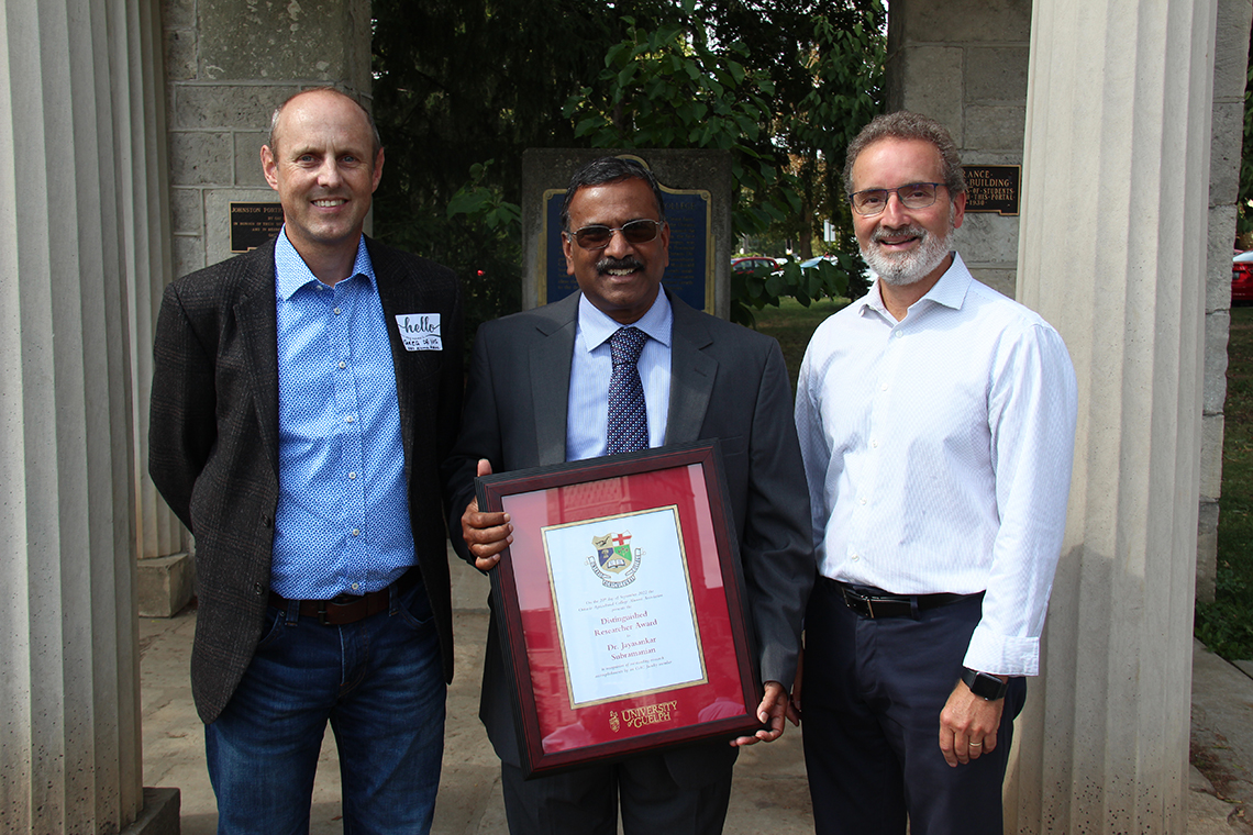 Three men in business casual clothing standing beside each other. The man in the center is holding an award.