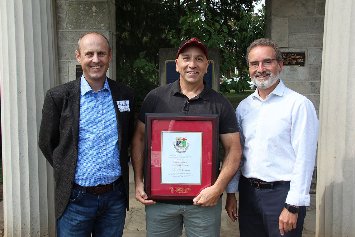 Three men in business casual clothing standing beside each other. The man in the center has a ball cap on and is holding an award.