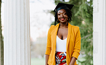 E'layna wearing a dress, yellow blazer and a graduate cap, standing under the Portico.