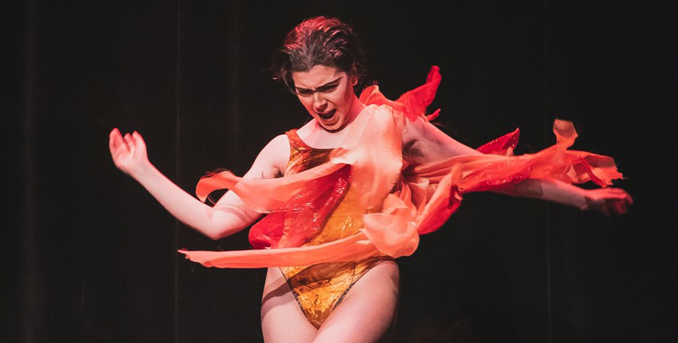 Big Mike in their climate change drag performance wearing an orange dress that resembles fire.