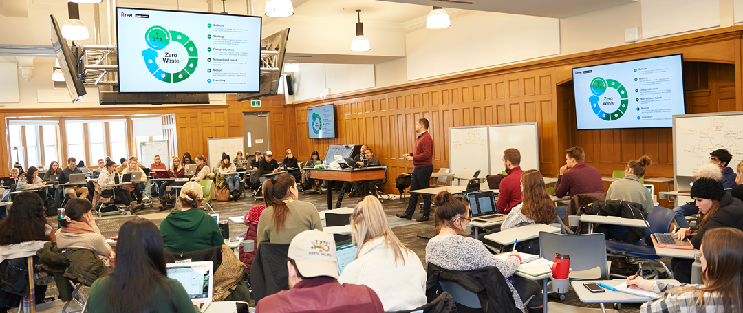 students in a classroom listening to a professor teach while tv's above the room present information