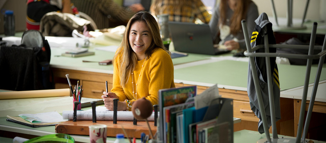 Student in yellow sweater smiles at camera while sitting at a drafting table with paper, books and computers around her