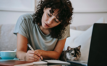 A student with long curly hair working at a desk with a cat on their lap..