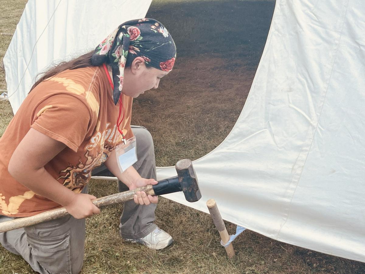 A person secures a teepee by hammering a wooden stake into the ground. 