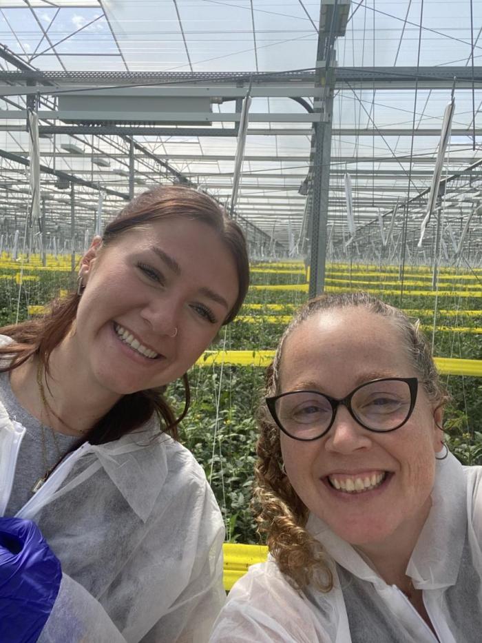 Two women stand in a greenhouse smiling.