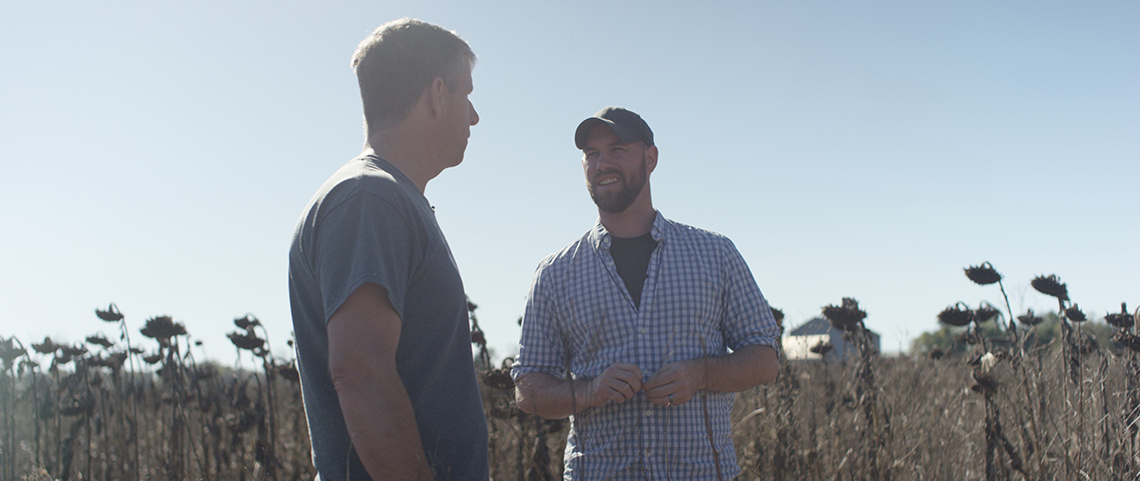 Two men talk while standing in a field. 