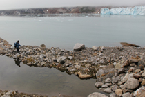 Hiker walks along rocks near open water, iceberg in background.