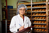 Food industry management student stands in a bakery with a clipboard.