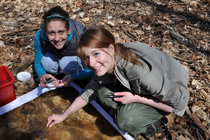 Two female students collect samples from a bin of water outside.