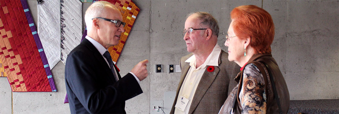 President Vaccarino speaks with the Roberts in a casual conversation. Grey walled room with colorful tapestry in the background.