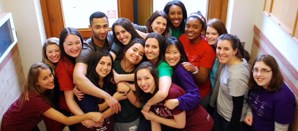 Group of male and female students stand together, arms around one another, in doorway