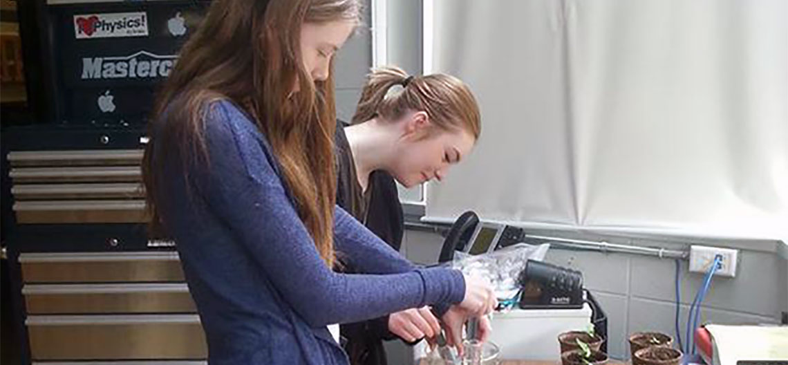 Two female high school students mixing fertilizer solution beside small sunflower pots
