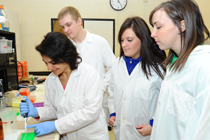 Three students in lab coats watch a fourth student use a pipette.
