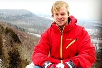 Jeremy sits on top of a snowy hill in a red jacket and mittens.