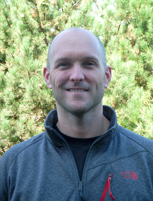 Ryan Prosser in grey sweater stands in front of green pine trees