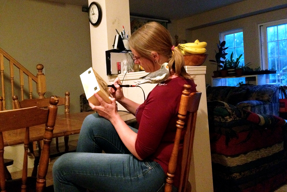 Sara Stricker works on a wood-burning art project as her pet bird sits on her shoulder.