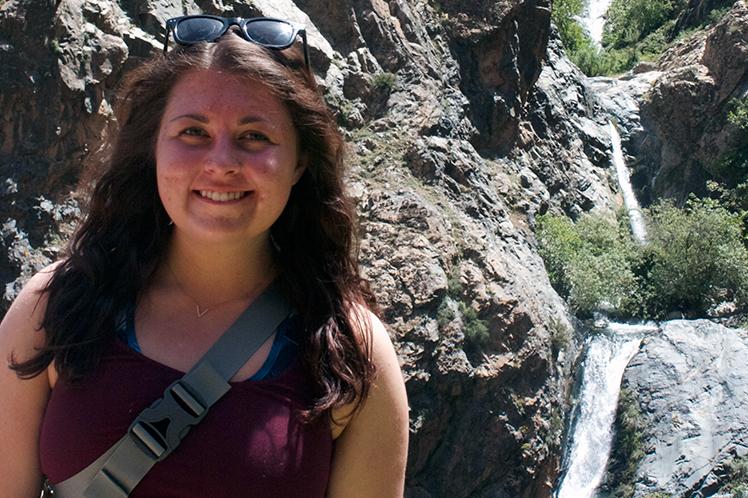 Laura Stortz stands in front of a rocky cliff with a small waterfall flowing through it.