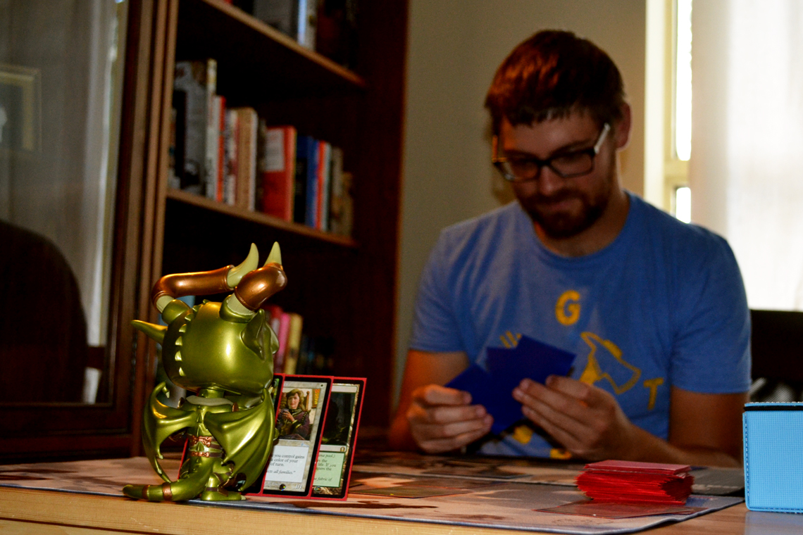 Zach sits across a table from a green figurine, both holding playing cards.