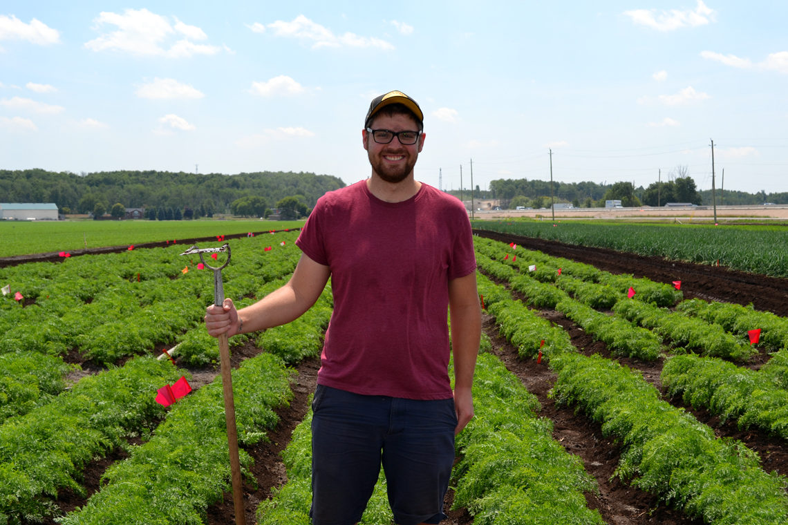 Zach stands in carrot field holding a pitchfork.