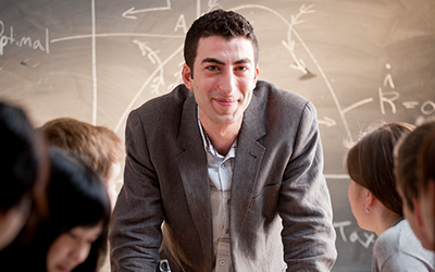 Instructor leaning on a table with students looking at them, in front of a blackboard with graphs and equations