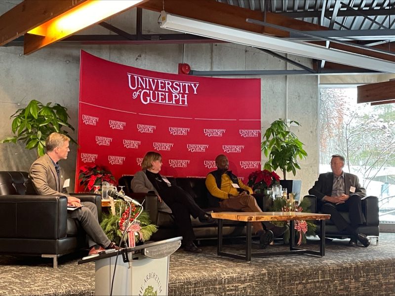 The panelists and modeator are seated on couches on a stage, mid-discussion. Behind them is a red banner with the University of Guelph logo. 