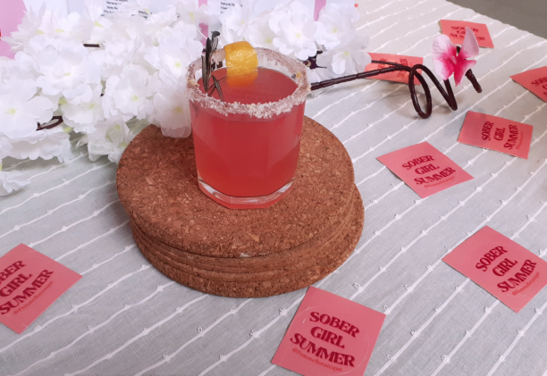 Salt-rimmed glass with pink cocktail displayed on cork coasters. Table has white flowers and pink stickers saying Sober Girl Summer scattered overtop.