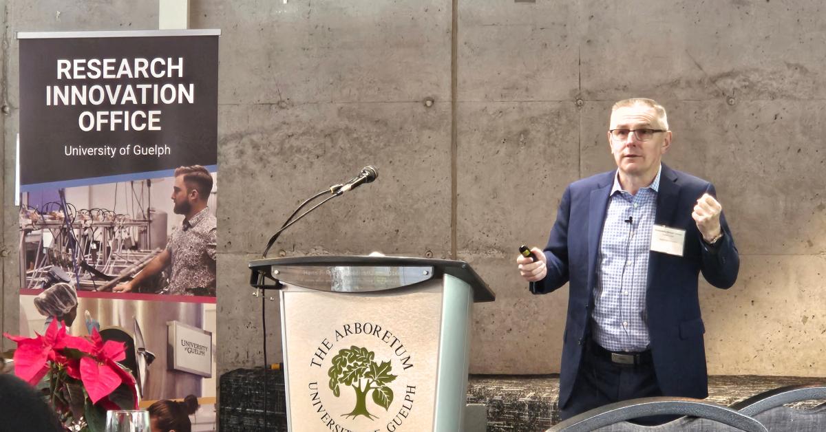 Justice Crinson stands beside a podium giving a presentation. A banner from the Research Innovation Office is on the other side of the podium. 