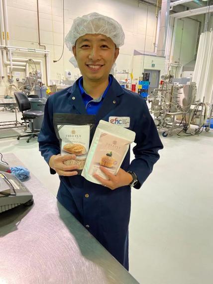 Dr Shawn Shao in the Guelph Food Innovation Centre, wearing navy blue coveralls and a hairnet, smiling and holding two bags of Treetly baking mixes. 