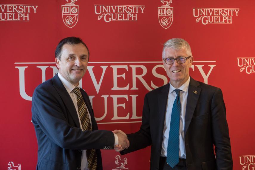 John Brennan and Malcolm Campbell shake hands in front of a UofG backdrop