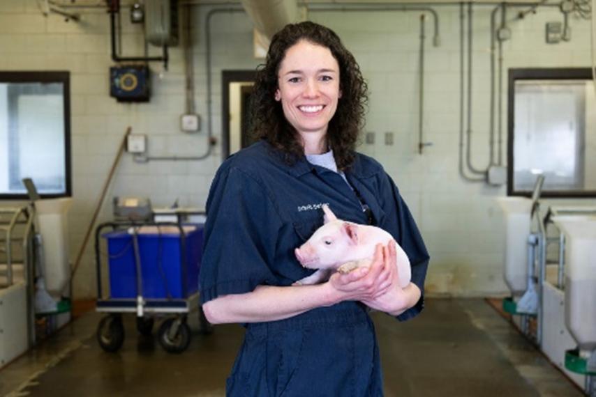 Dr Lee-Anne Huber smiling at the camera, wearing navy blue coveralls and cradling a pink baby piglet.