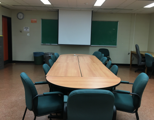 A white walled room containing a long table with several chairs and a projector screen