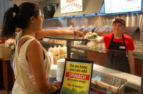 Hospitality Services' server Kendal West (right) delivers a wrap with added spinach to student Amia Khosla at the University Centre deli sandwich station. A sign beside Amia says "Did you know you can add spinach for no extra charge? Try it today!"