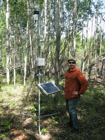 Prof. Gedalof stands in a forest beside a temporary weather station - a solar panel and wiring, mounted on a metal tripod