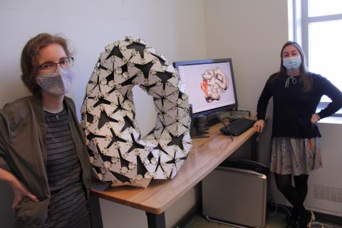 Diana Pfeffer (left), MLA student, stands beside a sculpture on a table. The sculpture is oval and black and white. Also on the table is a computer monitor.  In the right of the photo is Michael Richardson, an MFA history student. 