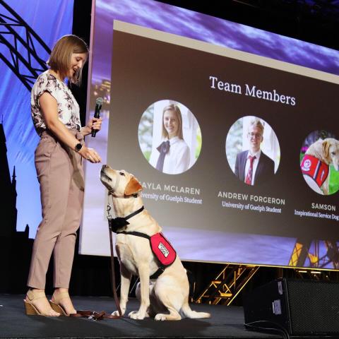 lady standing on the left with service dog sitting at her feet. both appear on a stage