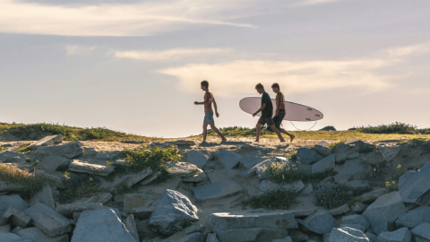 Three men walking along a rocky area with a surfboard