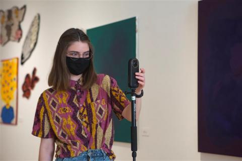 A woman wearing a mask and standing in an art gallery. She is touching a camera on a tripod with her left hand. 