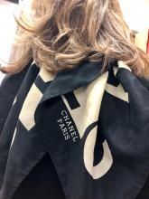 A woman's back is to the camera.  Her hair is brown, and shoulder length.  She wears a black sweater and a black and white scarf that says Chanel, Paris on it.