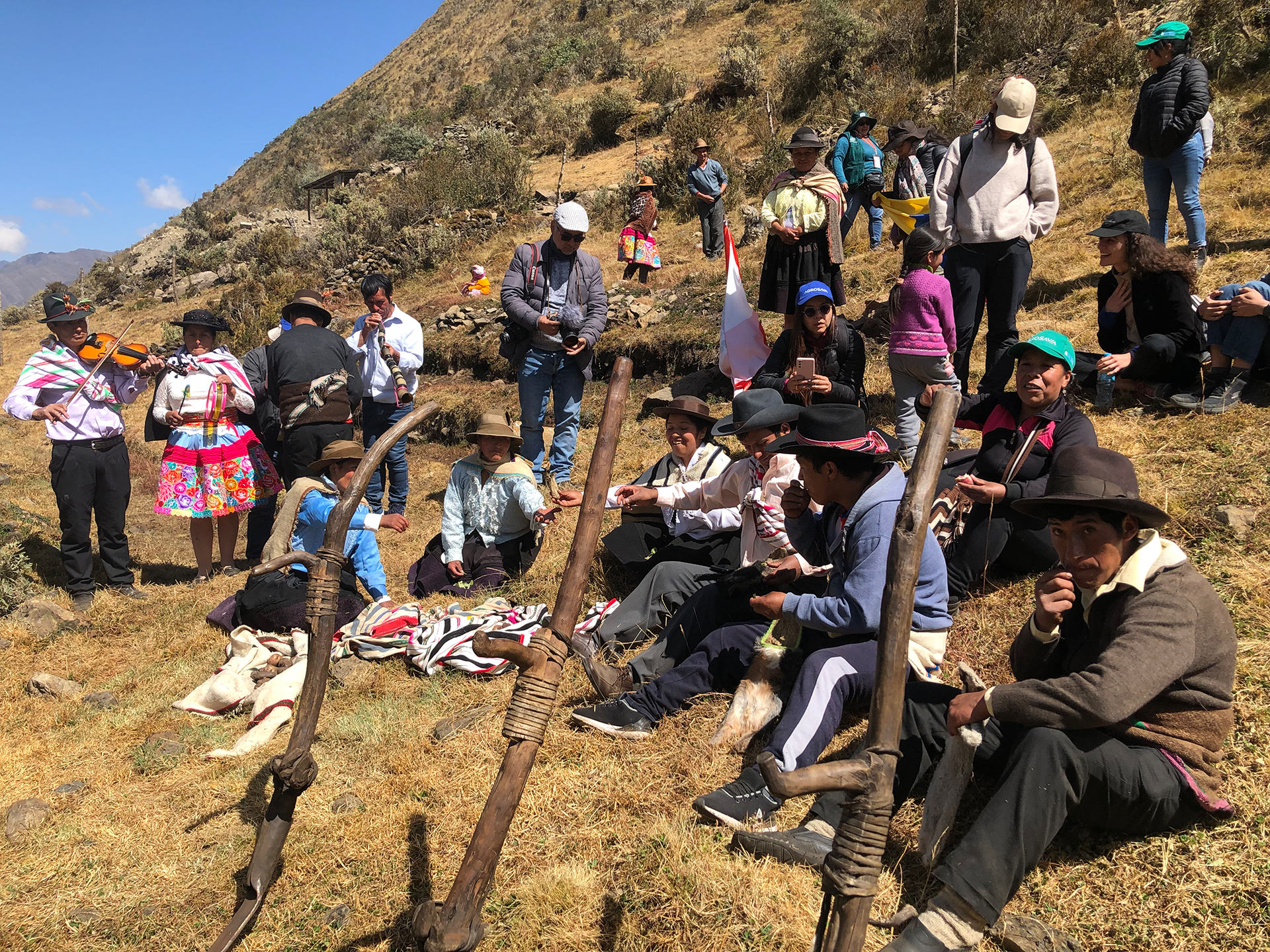 A group of people gathering on the side of the mountain. Some are sitting while others are standing. A few people have musical instruments including a horn, tambourine and violin.