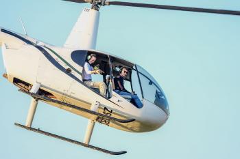 A white helicopter flies against a clear blue sky while a researcher wearing a headset leans out of the open doorway operating darting equipment. The pilot sits beside them as the helicopter conducts wildlife research over the South African landscape.