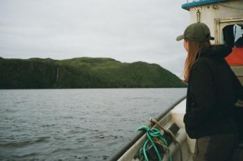 A woman standing on a boat looking out towards water and a mountainous landscape.