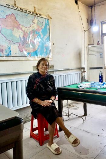 Inside the break room of a Chinese farmer, the farmer sits on a red plastic stool at her Mahjong play table. In the background, an old Chinese flag at the wall and a single bright light bulb.